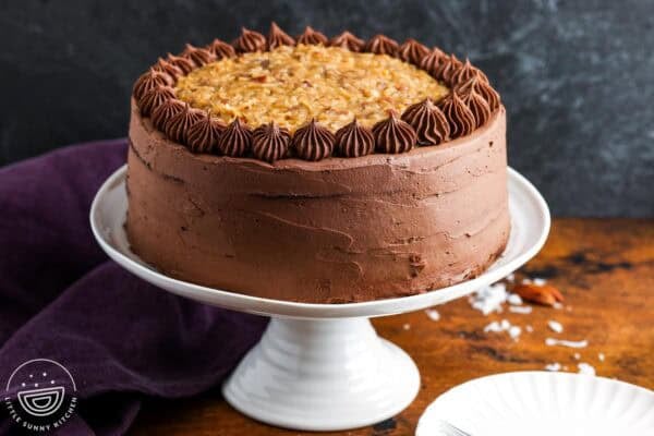 A german chocolate layer cake on a white cake stand set on a wooden table next to a dark purple towel.