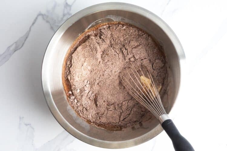 dry ingredients added to wet ingredients to make a german chocolate cake batter in a metal bowl with a whisk.