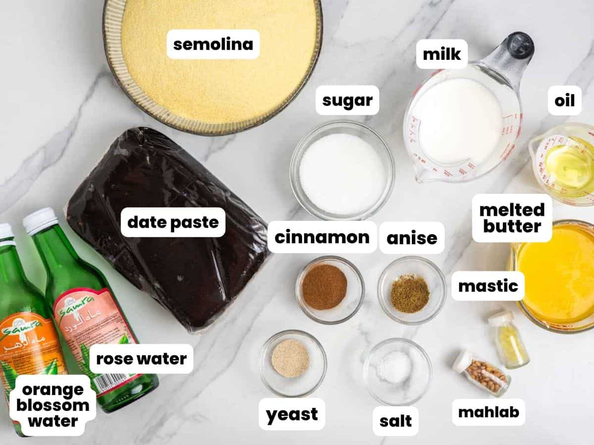 A flat lay of labeled baking ingredients including semolina, date paste, milk, sugar, oil, melted butter, rose water, orange blossom water, cinnamon, anise, yeast, salt, mastic, and mahlab on a light marble surface.