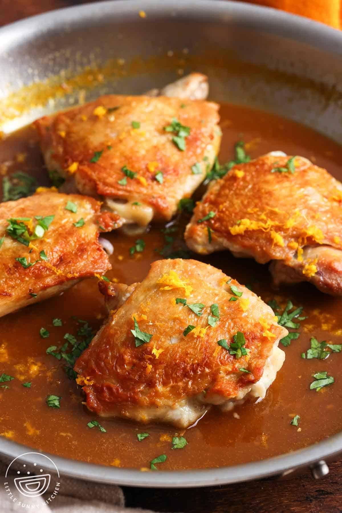 crispy chicken thighs in a skillet with orange sauce, garnished with orange zest and chopped parsley.
