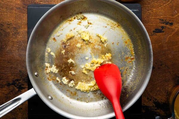 minced garlic cooking in a stainless steel skillet, stirred with a red spoon.