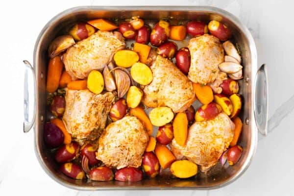 raw bone-in, skin-on chicken thighs in a roasting pan with quartered red onion, halved baby potatoes, and sliced carrots.