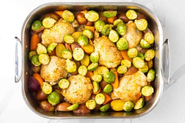 Seasoned, partially baked chicken thighs in a roasting pan, surrounded by halved brussels sprouts, baby potatoes, and sliced carrots.