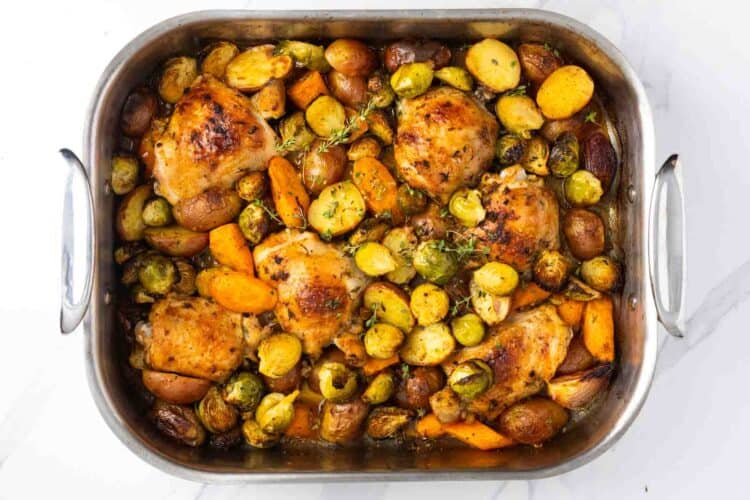overhead view of a large metal roasting pan filled with roasted chicken thighs and roasted vegetables, including potatoes, carrots, and brussels sprouts.