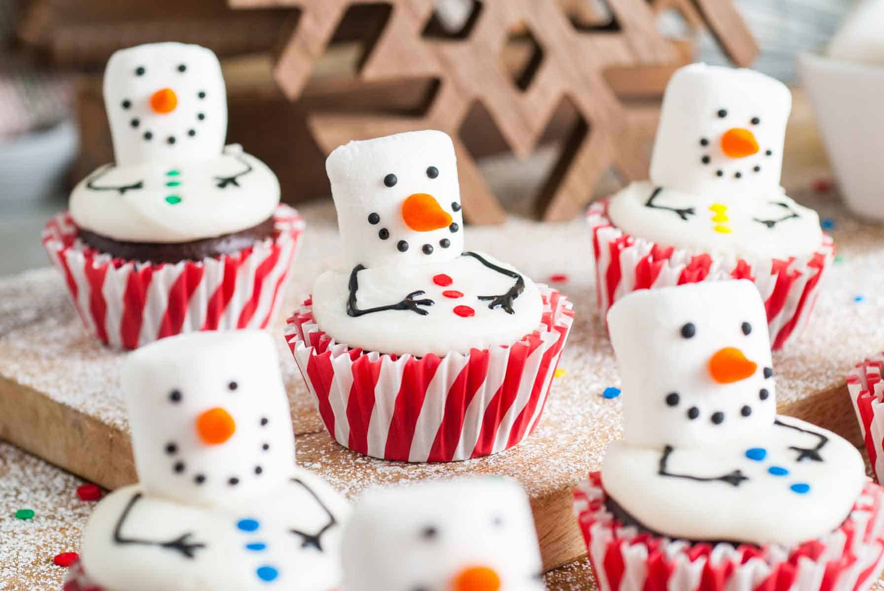Chocolate cupcakes topped with white fondant snowmen featuring marshmallow heads carrot noses and button details on a festive holiday setup