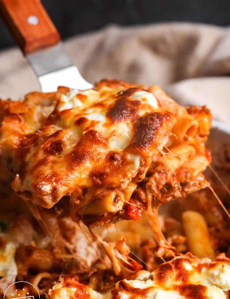 a square of baked ziti lifted out of the pan with a spatula to show the meaty, saucy pasta and lots of melted cheese.