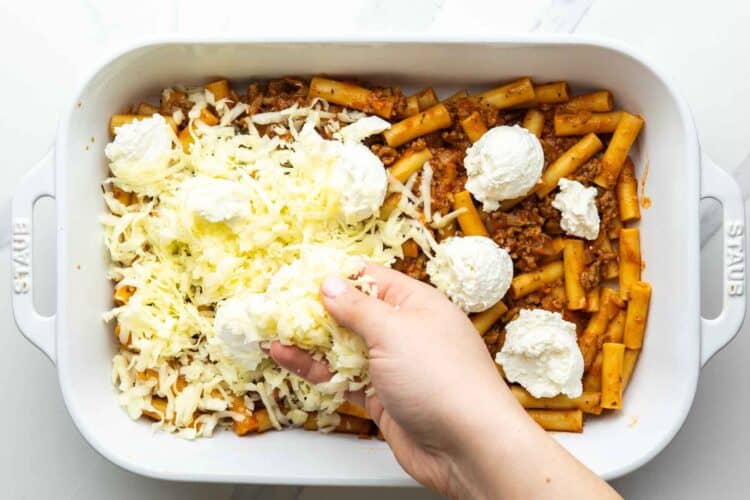 a hand adding dollops of ricotta cheese to the top of a pan of baked ziti, then adding shredded mozzarella cheese over that.