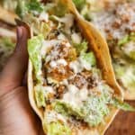 Hand holding a folded Caesar chicken smash taco showing crispy tortilla, seasoned chicken, romaine lettuce, Parmesan cheese, and Caesar dressing inside.