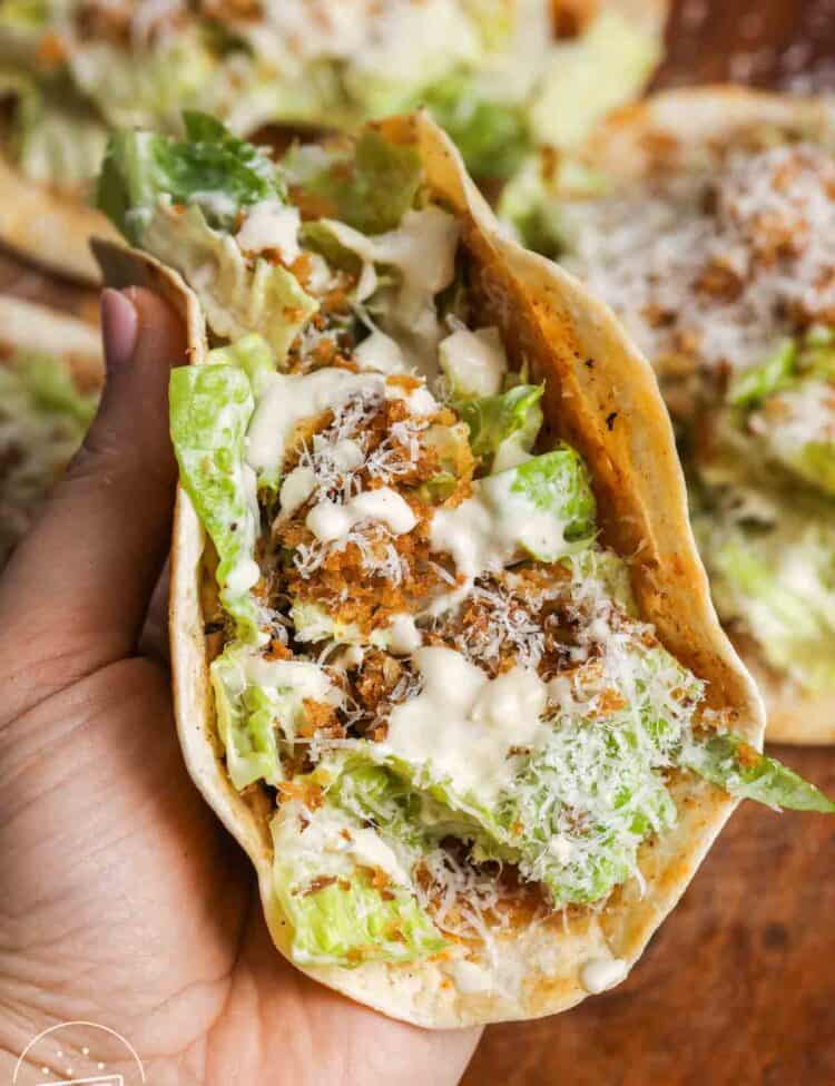 Hand holding a folded Caesar chicken smash taco showing crispy tortilla, seasoned chicken, romaine lettuce, Parmesan cheese, and Caesar dressing inside.
