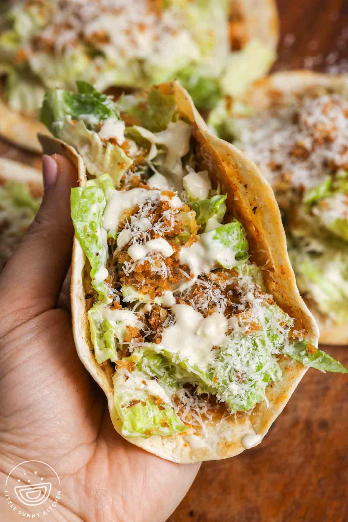 Hand holding a folded Caesar chicken smash taco showing crispy tortilla, seasoned chicken, romaine lettuce, Parmesan cheese, and Caesar dressing inside.