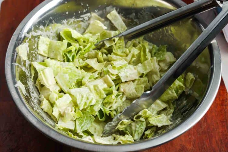 Romaine lettuce tossed with Caesar dressing in a stainless steel bowl with tongs.
