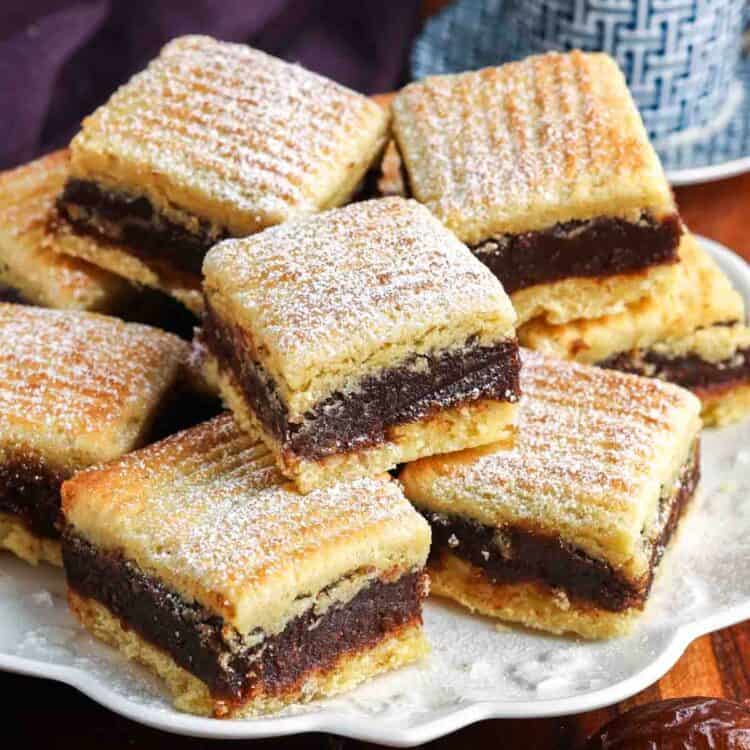 Stacked maamoul semolina date bars dusted with powdered sugar on a serving plate, with a cup of coffee in the background.