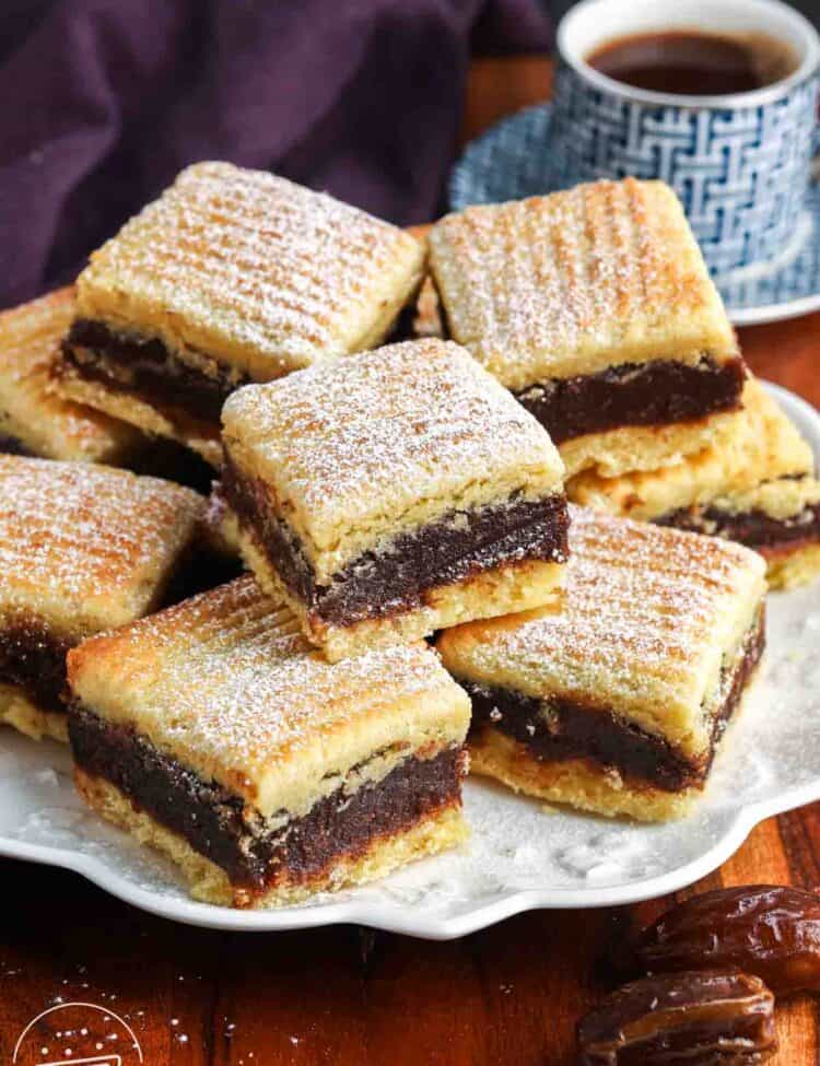 Stacked maamoul semolina date bars dusted with powdered sugar on a serving plate, with a cup of coffee in the background.