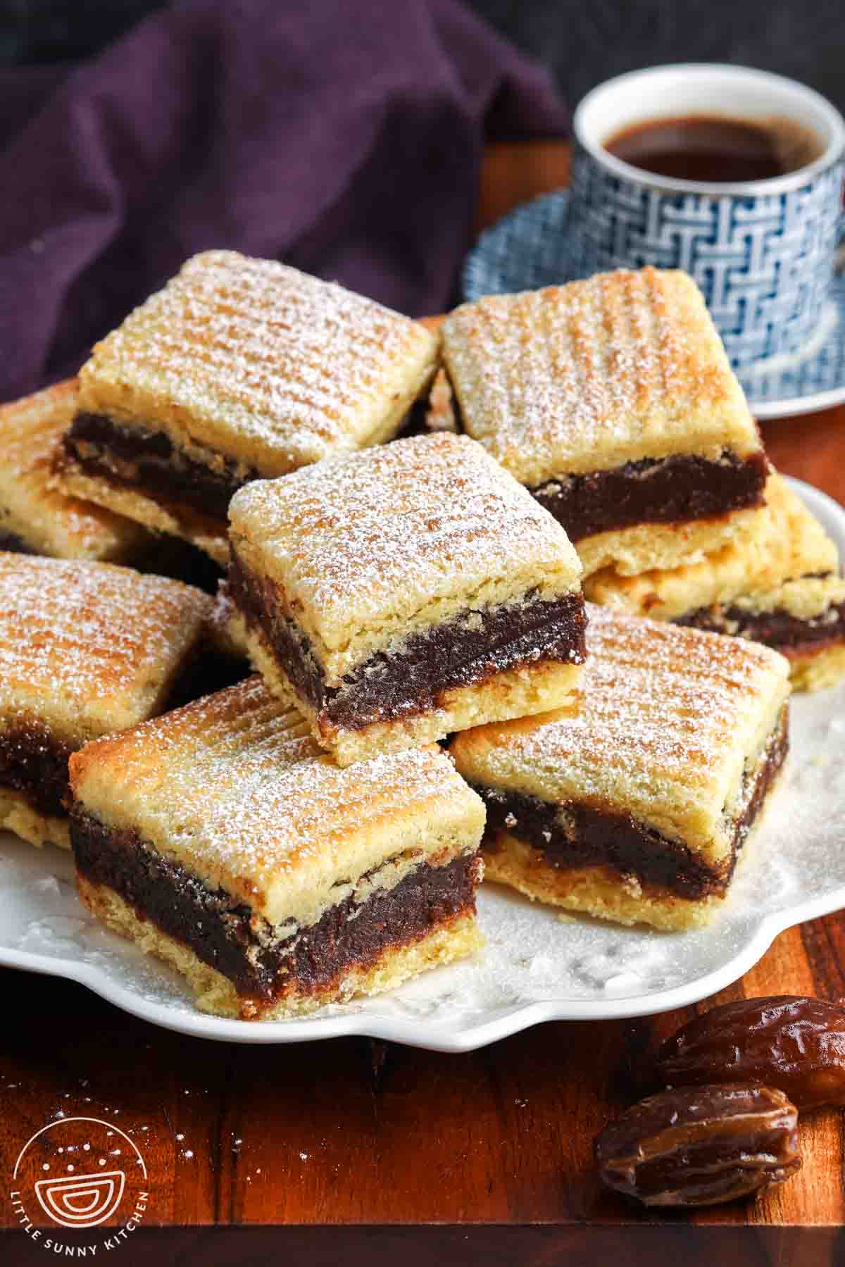 Stacked maamoul semolina date bars dusted with powdered sugar on a serving plate, with a cup of coffee in the background.