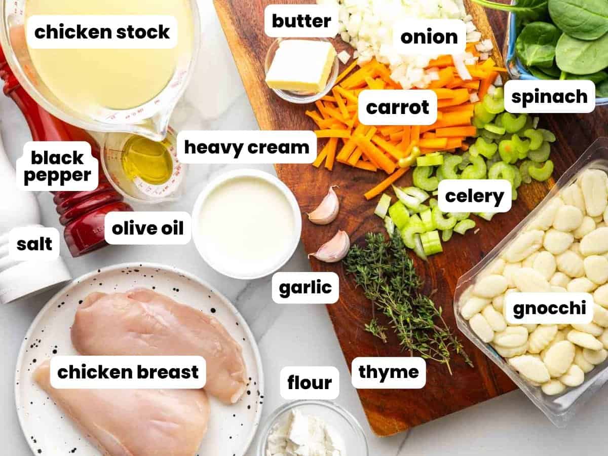 The ingredients needed to make an olive garden copycat chicken gnocchi soup, including veggies, chicken breast, gnocchi, fresh thyme, garlic cloves, and chicken stock. The ingredients are on a wooden cutting board set on a marble counter.