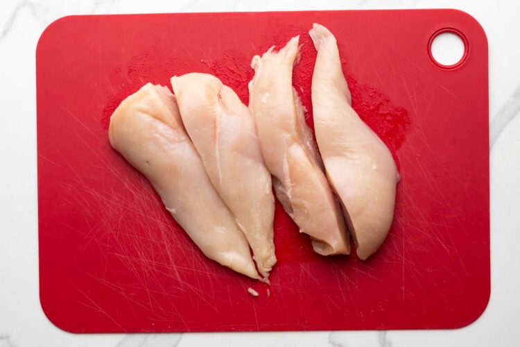 Two chicken breasts, cut in half lengthwise on a red plastic cutting board.