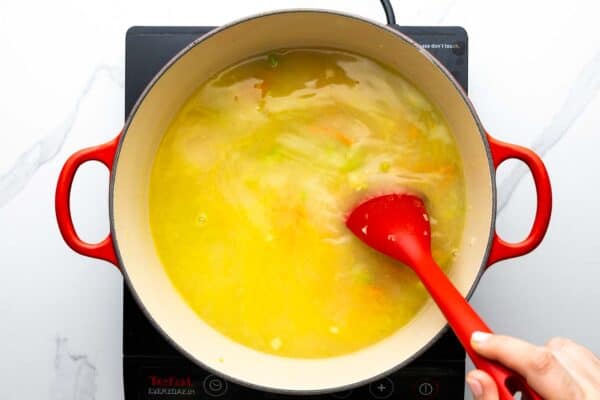 Chicken broth added to soup in a dutch oven, stirred with a red spoon.