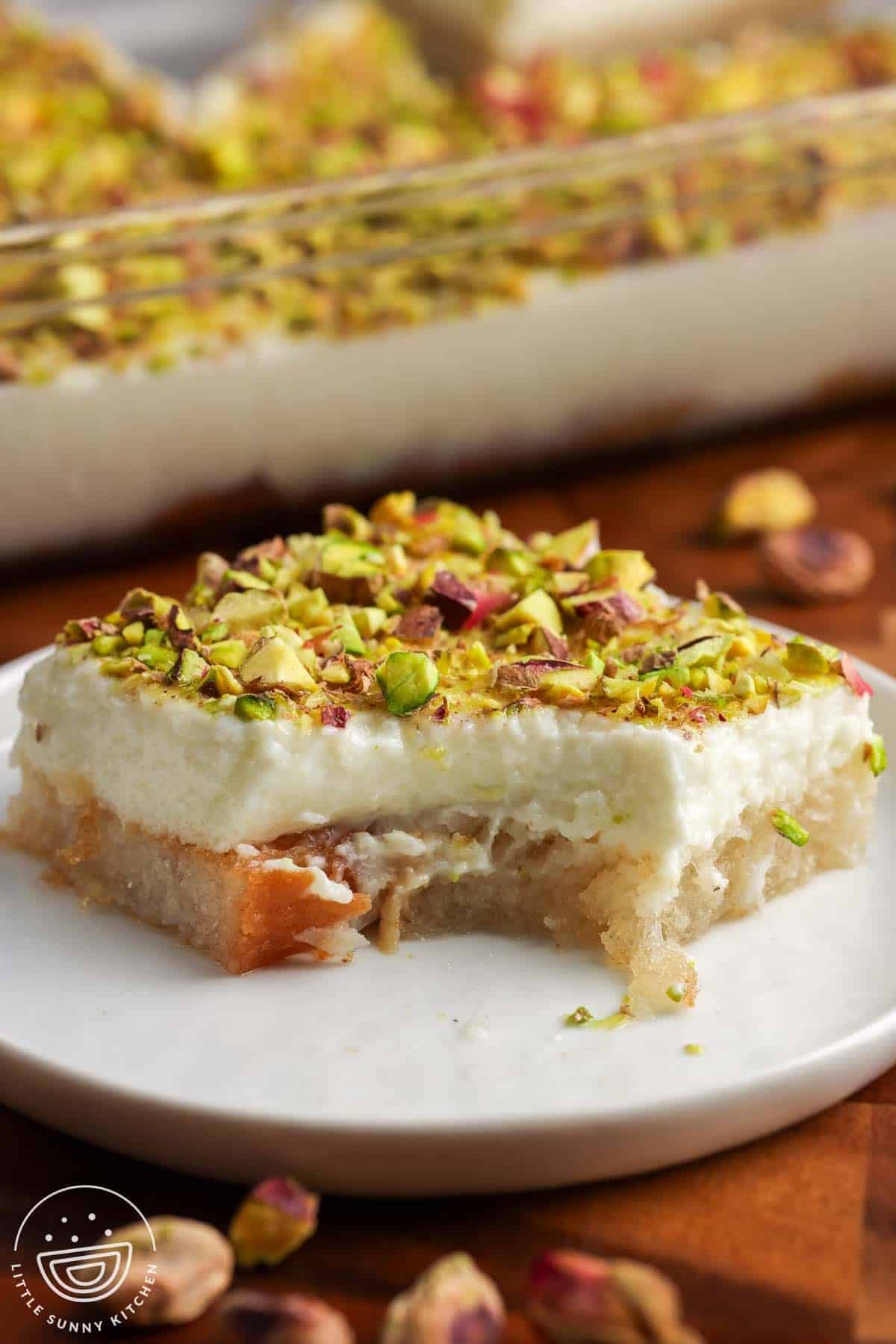 Square slice of Aish El Saraya topped with chopped pistachios, showing layers of syrup soaked toasted bread and thick ashta cream, served on a white plate with the remaining dessert in a glass pan in the background.