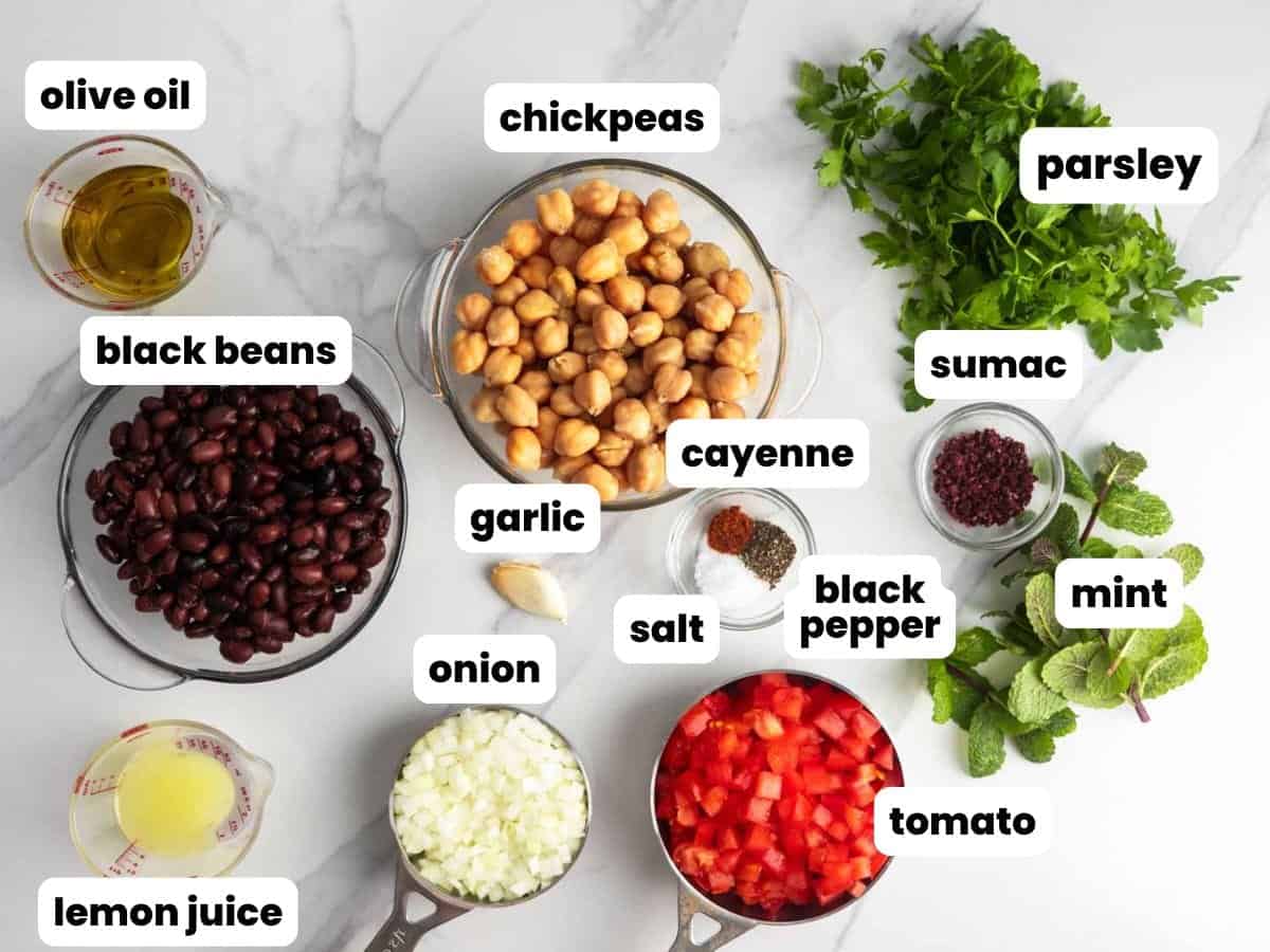 Overhead image of balela salad ingredients arranged in bowls on a marble surface, including chickpeas, black beans, diced tomatoes, diced onion, fresh parsley, fresh mint, garlic, lemon juice, extra virgin olive oil, salt, black pepper, cayenne, and sumac, each labeled.