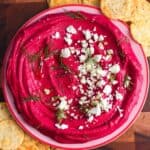 Overhead photo of creamy beet hummus swirled in a shallow bowl, topped with crumbled feta and fresh dill, served with round crackers on a wooden board.