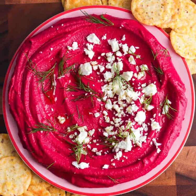 Overhead photo of creamy beet hummus swirled in a shallow bowl, topped with crumbled feta and fresh dill, served with round crackers on a wooden board.