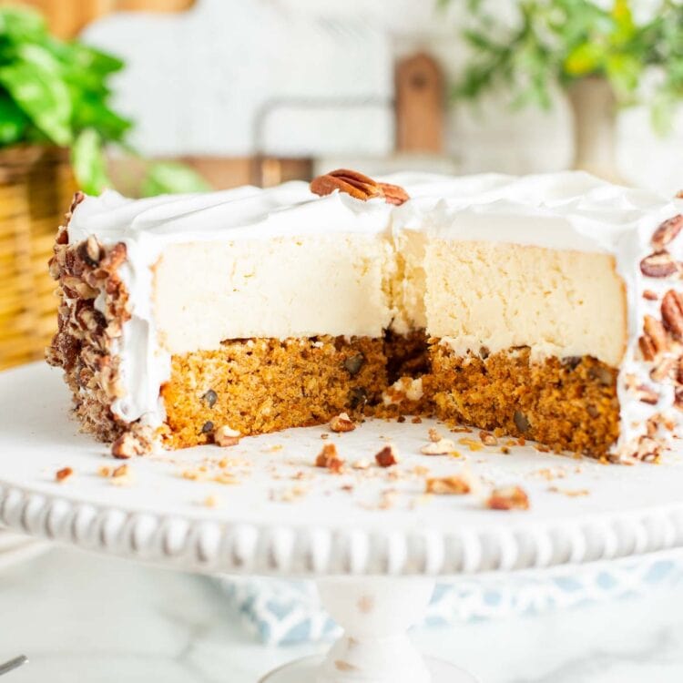 double layered carrot cake cheesecake on a pedestal, with slices removed. the cake is covered in whipped cream and then with chopped pecans.