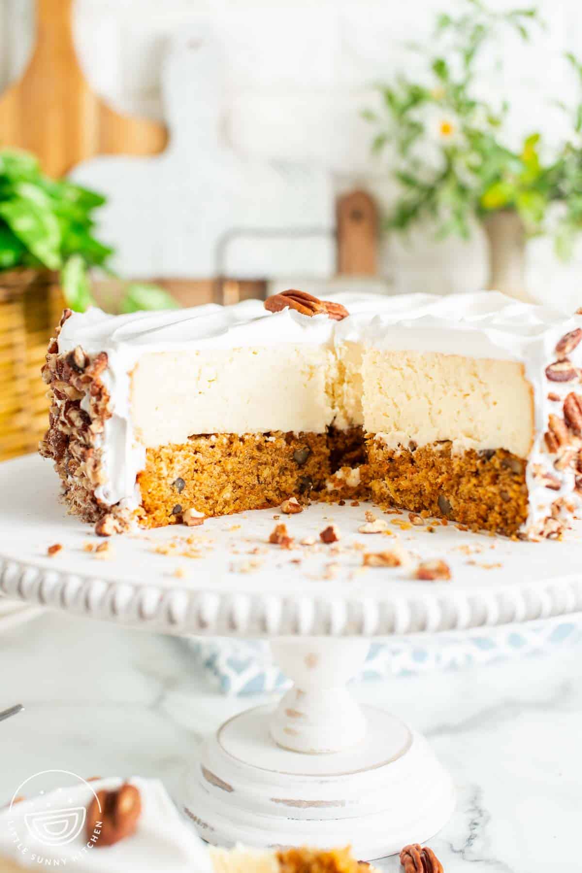 double layered carrot cake cheesecake on a pedestal, with slices removed. the cake is covered in whipped cream and then with chopped pecans.