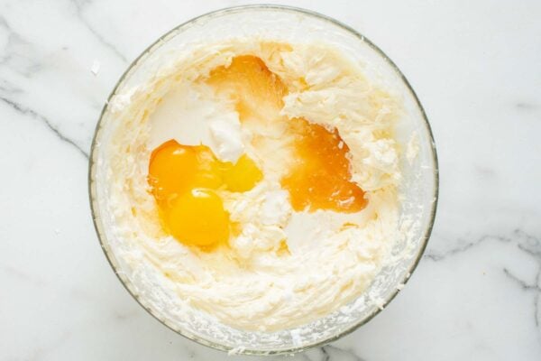 Eggs and vanilla added to whipped cream cheese in a large glass mixing bowl.
