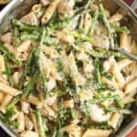 a skillet of creamy penne with asparagus and diced chicken mixed in. The skillet is set on a wooden board.