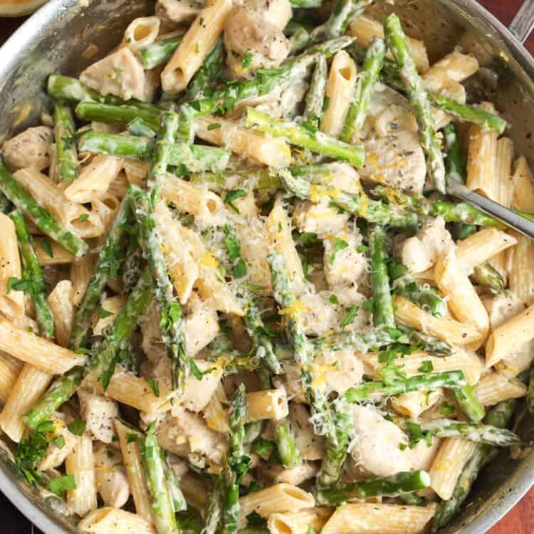 a skillet of creamy penne with asparagus and diced chicken mixed in. The skillet is set on a wooden board.