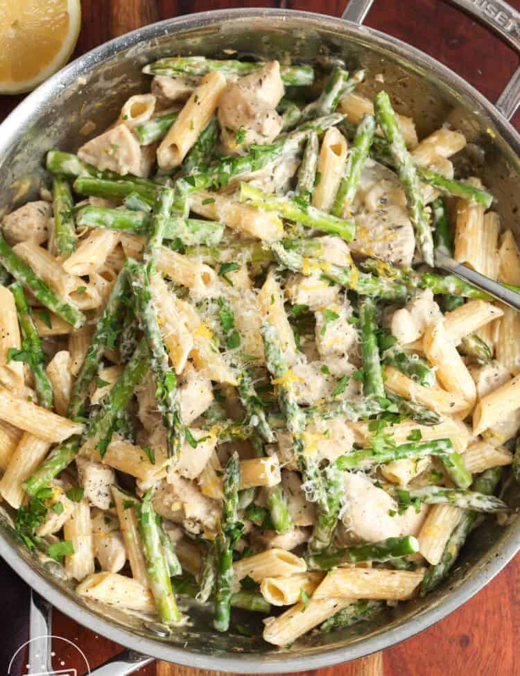 a skillet of creamy penne with asparagus and diced chicken mixed in. The skillet is set on a wooden board.