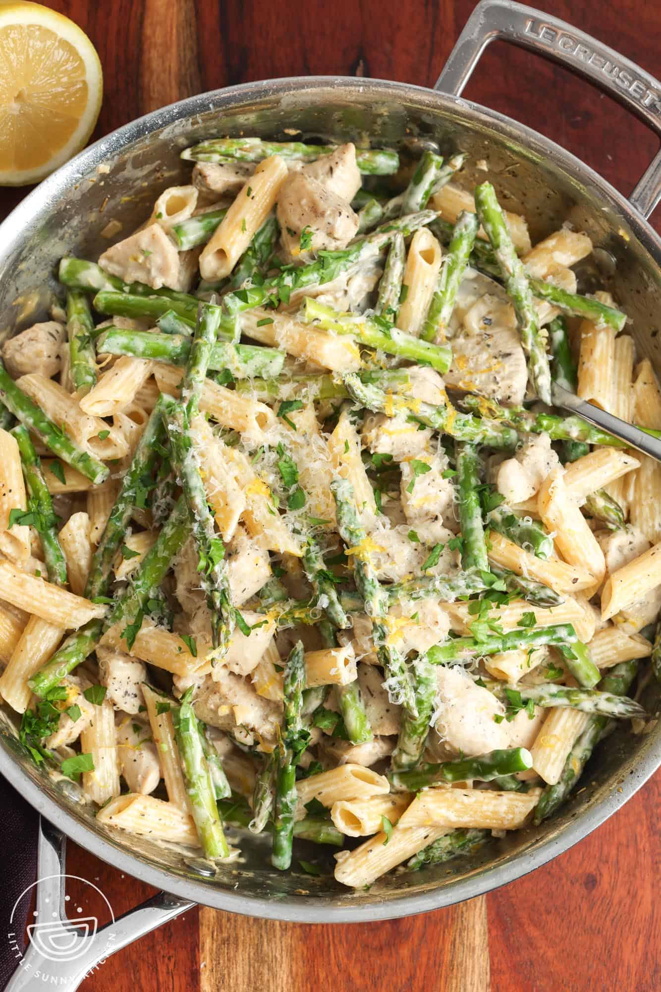 a skillet of creamy penne with asparagus and diced chicken mixed in. The skillet is set on a wooden board.