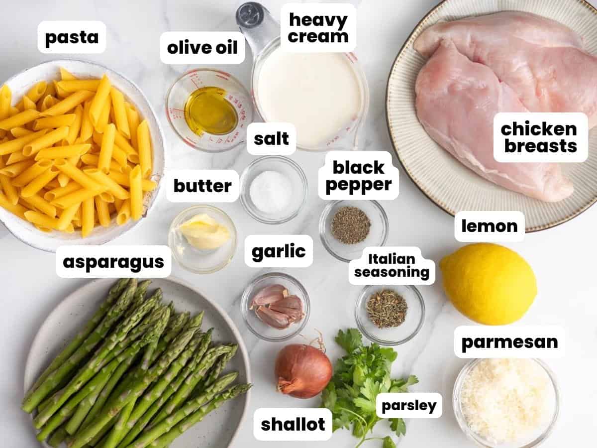 Plates holding raw chicken breasts, penne pasta, and asparagus spears surround small bowls holding the rest of the ingredients needed to make a creamy asparagus and chicken pasta dish. All of the ingredients are arranged on a marble countertop.