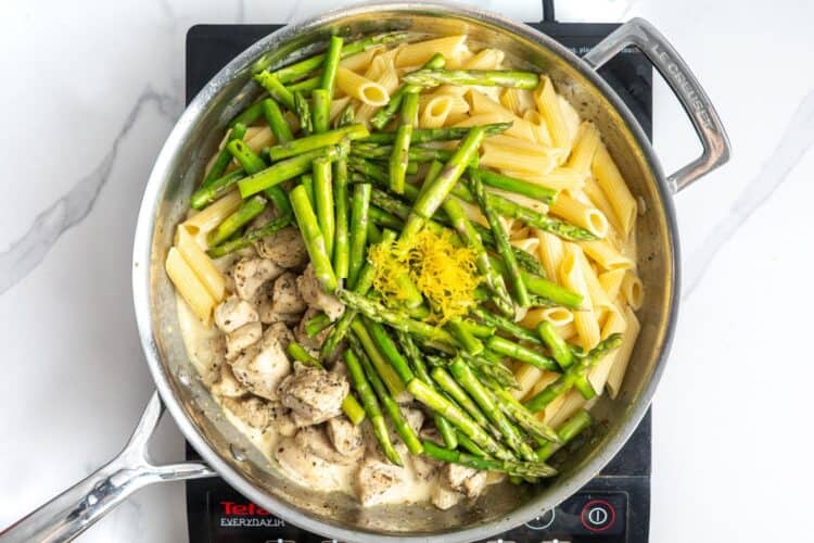 cooked pasta, cooked chicken, and cooked asparagus added to creamy sauce in a skillet. Lemon zest is on top of the asparagus.