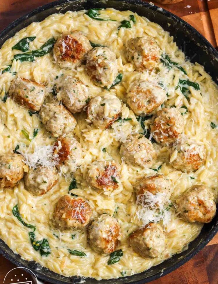 a frying pan of creamy orzo pasta with spinach and chicken meatballs, set on a wooden cutting board, garnished with parmesan cheese.