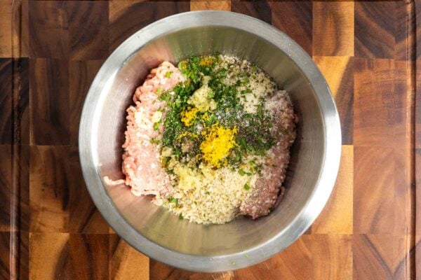 A pound of ground chicken in a metal bowl on a wooden cutting board. On top of the meat are breadcrumbs, garlic, parsley, lemon zest, salt, and pepper.