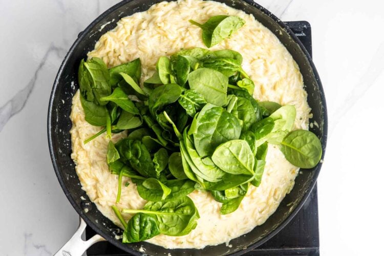 a handful of baby spinach added to creamy orzo pasta in a skillet.