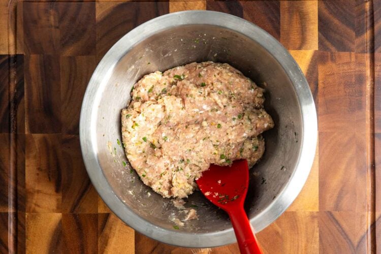 Raw meat mixture for lemon chicken meatballs in a metal mixing bowl set on a wooden cutting board.