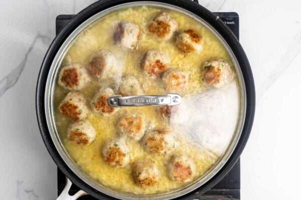 Orzo and chicken meatballs cooking in s skillet, covered with a lid