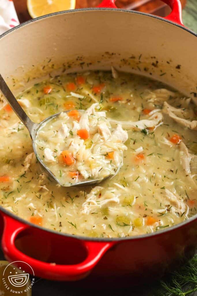 A red Dutch oven filled with lemon chicken orzo soup, featuring shredded chicken, tender orzo pasta, diced carrots, celery, and fresh dill, with a ladle scooping up a hearty portion.
