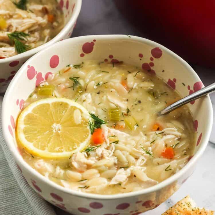 A bowl of lemon chicken orzo soup topped with a fresh lemon slice, filled with shredded chicken, orzo, carrots, celery, and dill, served with a spoon and a piece of garlic bread on the side, with a red pot in the background.