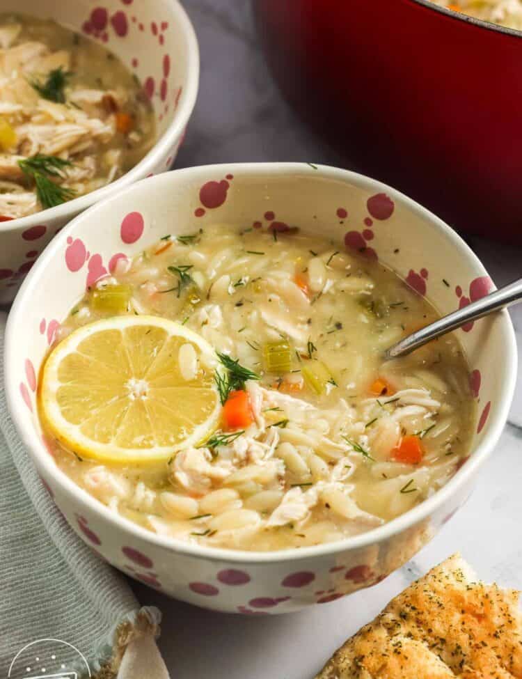 A bowl of lemon chicken orzo soup topped with a fresh lemon slice, filled with shredded chicken, orzo, carrots, celery, and dill, served with a spoon and a piece of garlic bread on the side, with a red pot in the background.
