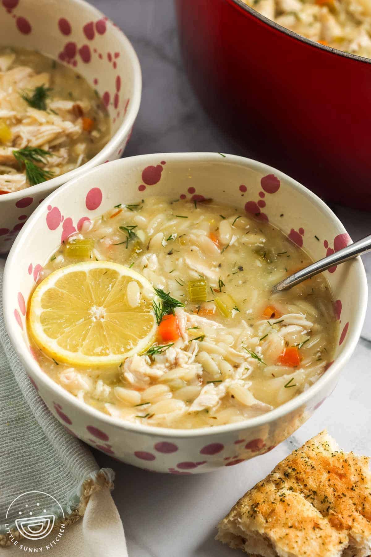 A bowl of lemon chicken orzo soup topped with a fresh lemon slice, filled with shredded chicken, orzo, carrots, celery, and dill, served with a spoon and a piece of garlic bread on the side, with a red pot in the background.