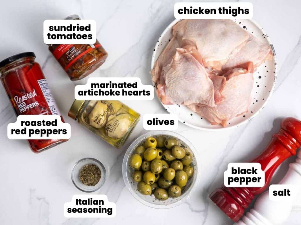 A plate of raw bone-in chicken thighs next to a jar of sundried tomatoes, a jar of roasted red peppers, a plastic container of olives, a jar of artichoke hearts, a small bowl of italian seasoning, and salt and pepper mills.