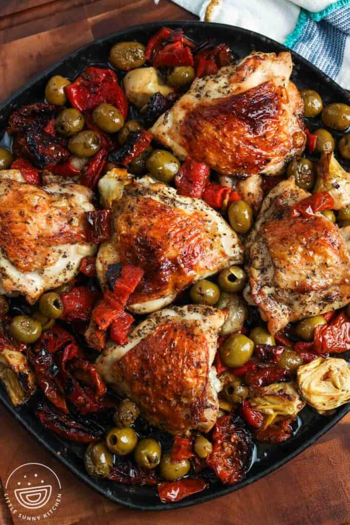 Crispy bone in chicken thighs baked with olives and red peppers, on a black rectangular platter.