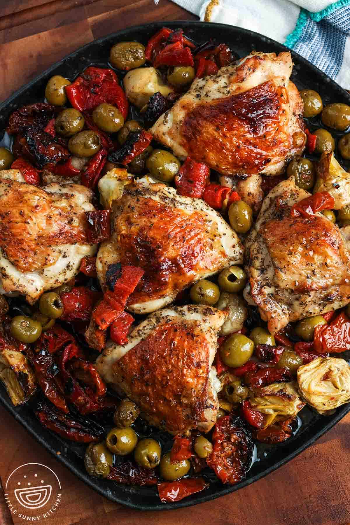 Crispy bone in chicken thighs baked with olives and red peppers, on a black rectangular platter.