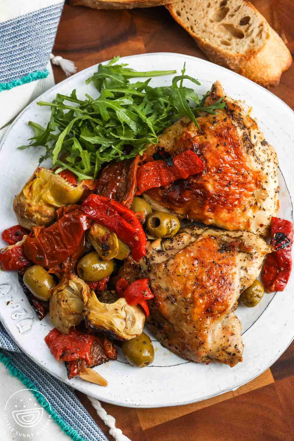 a white dinner plate of baked chicken, roasted olives and veggies, and green salad.
