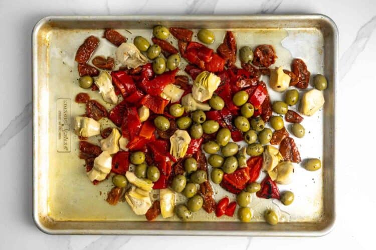 Olives, sundried tomatoes, artichokes, and red peppers with oil on a metal sheet pan.