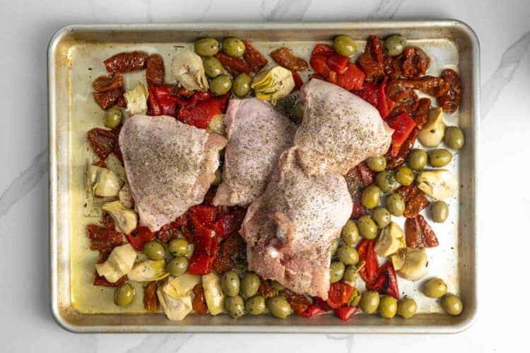 raw chicken thighs on top of olive mix on a metal sheet pan.