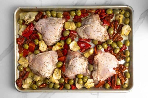 six chicken thighs arranged on a sheet pan, surrounded by olives, artichokes, and roasted red pepper strips.
